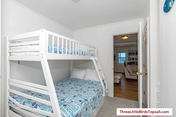 A bunk bed with a patterned bedspread, a door leading to a living area with a sofa and a coffee table, and a bookshelf in the background.
