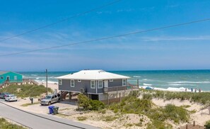 Une maison de plage avec un balcon donnant sur l’océan, une route au premier plan et un ciel dégagé.