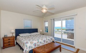 Une chambre à coucher comprenant un lit, une table de chevet, un banc, un ventilateur de plafond et une grande porte-fenêtre coulissante donnant sur un balcon.