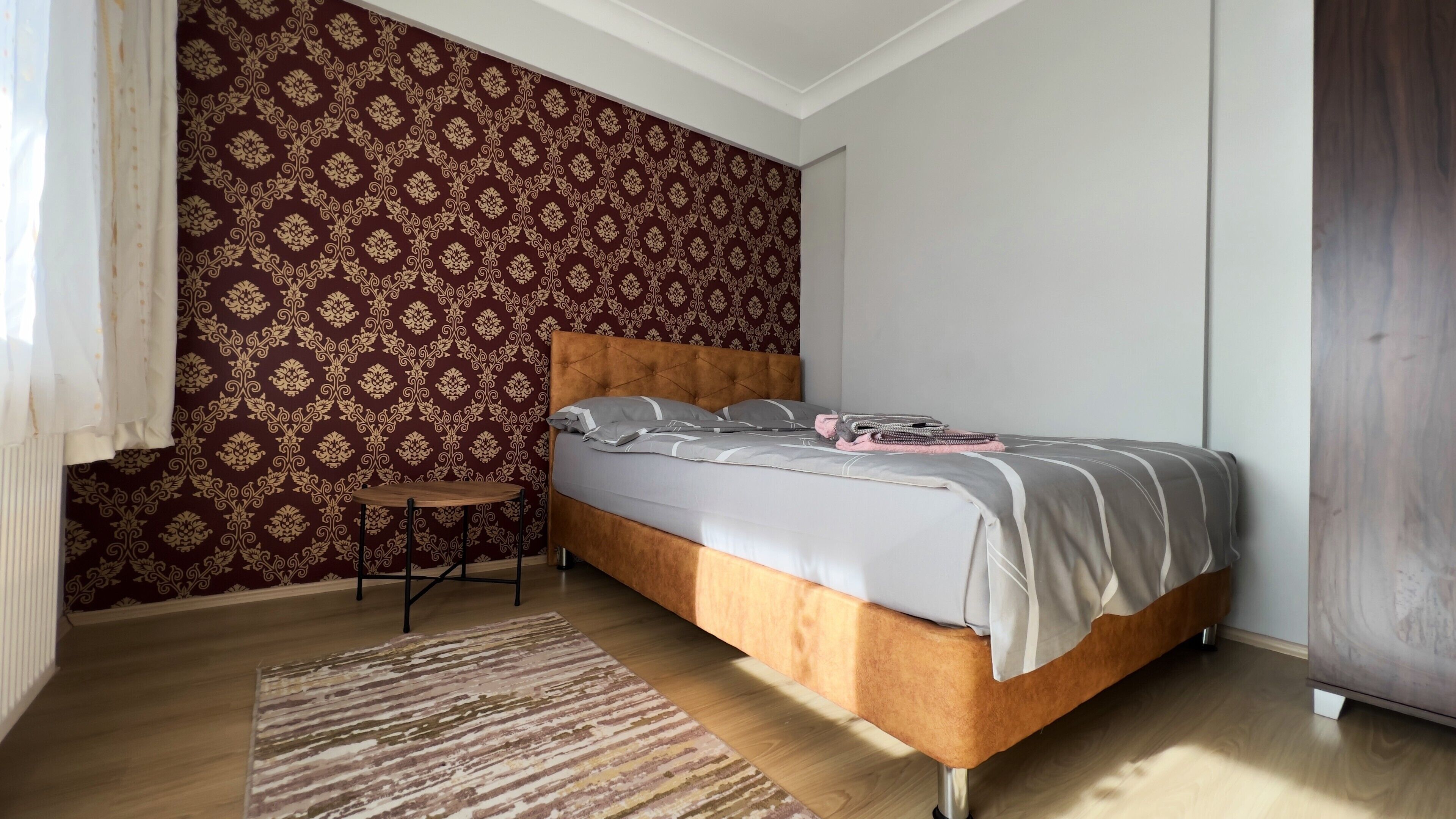 A bedroom with a patterned wallpaper, a bed with grey bedding, a small table, and a striped rug.
