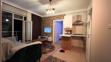 A compact hotel room with a bed, a small desk, a television, a kitchen area with a stove and refrigerator, and a stone accent wall.