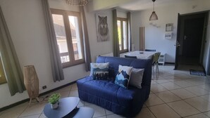 Een woonkamer met een blauwe bank, een bijzettafeltje met een plant en een eethoek met een tafel en stoelen.