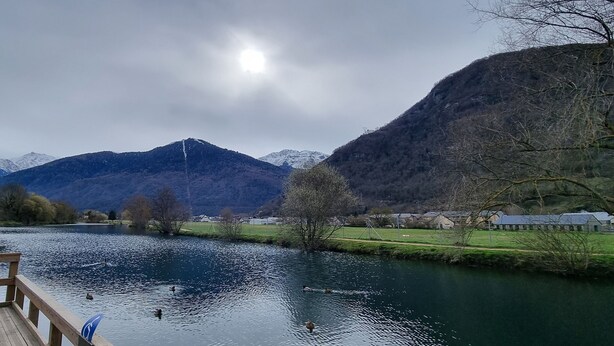 Een houten terras met uitzicht op een rivier, waar eenden zwemmen, omringd door bergen en een bewolkte lucht.