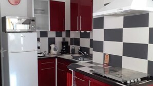 Een kleine keuken met rode kastjes, een witte koelkast, een zwart-wit geruit spatwand, en een centraal eiland met een kookplaat.