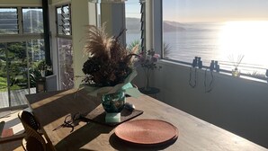 En spiseplads med et træbord, en vase med tørrede planter og havudsigt gennem store vinduer.