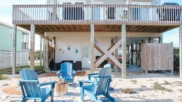 Une maison de plage dotée d’une terrasse couverte, d’un espace extérieur aménagé avec des fauteuils Adirondack et d’une clôture en bois.