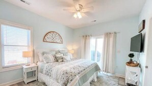 Une chambre à coucher comprenant un lit, des tables de chevet, un ventilateur de plafond, une fenêtre avec des stores et un téléviseur fixé au mur.