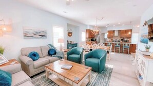 Un salon moderne avec un canapé, une table basse et des fauteuils de couleur turquoise. On aperçoit à l’arrière-plan une cuisine avec des meubles en bois et un coin repas.