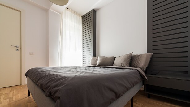 A modern bedroom with a bed, wooden flooring, a window with curtains, and a dark wall panel.