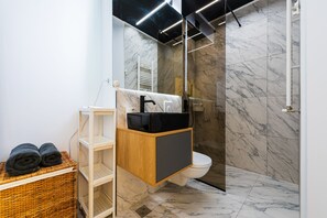 Un bagno moderno con pavimento in marmo, un mobile bagno in legno, un cesto di vimini con asciugamani e una cabina doccia in vetro.
