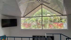 A room with a large window, a ceiling fan, a flat-screen TV, a bed, a staircase, and a Buddha head sculpture. - Casas Guaney (Quepos)