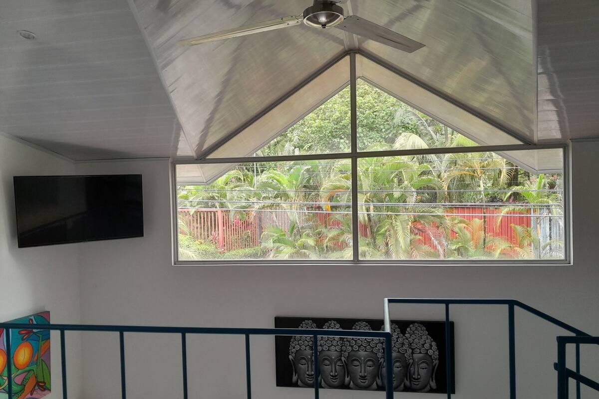 Una habitación con un ventanal grande, un ventilador de techo, un televisor de pantalla plana, una cama, una escalera y una escultura de cabeza de Buda.