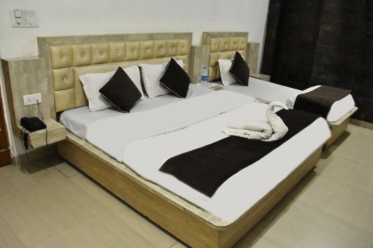 Uma cama de casal com cabeceira bege, roupa de cama branca e almofadas pretas. Há uma mesinha de cabeceira com um telefone e uma garrafa de água.