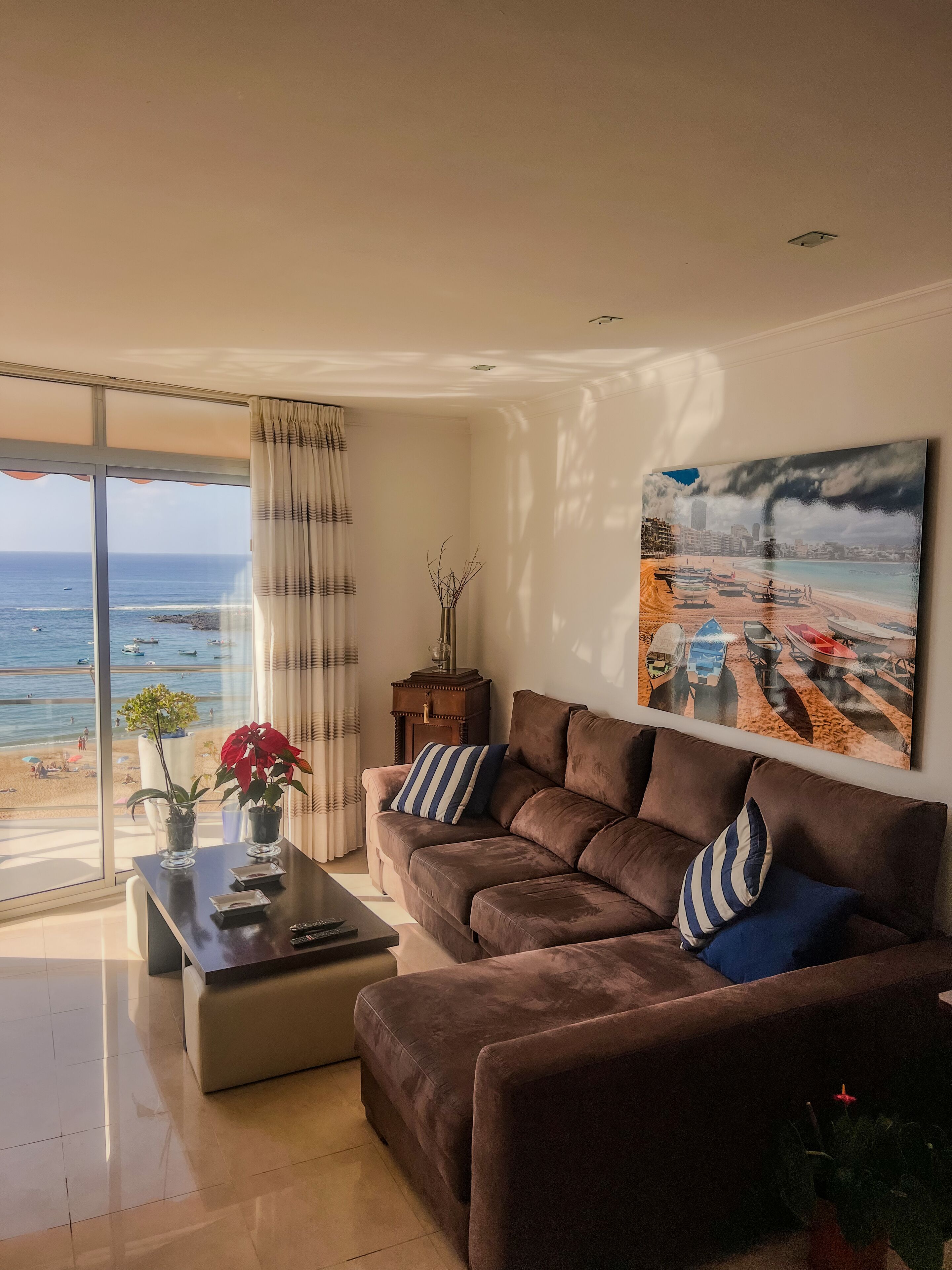 Una sala moderna con un gran ventanal que da a una playa, un sofá marrón con cojines a rayas, una mesita con un jarrón de flores y un cuadro de una escena playera en la pared.