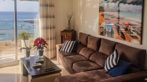 Un soggiorno moderno con un'ampia finestra vista spiaggia, un divano marrone con cuscini a righe, un tavolino con un vaso di fiori e un quadro raffigurante una spiaggia.
