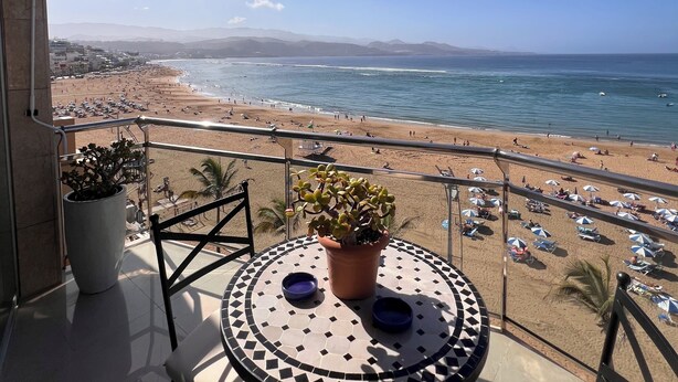 Un balcone con vista sulla spiaggia e sull'oceano. Si nota un tavolo rotondo con sedie e una pianta in vaso.