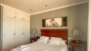 Un letto rifatto con lenzuola e asciugamani bianchi, una testiera in legno, un comodino con una lampada e un quadro appeso al muro.
