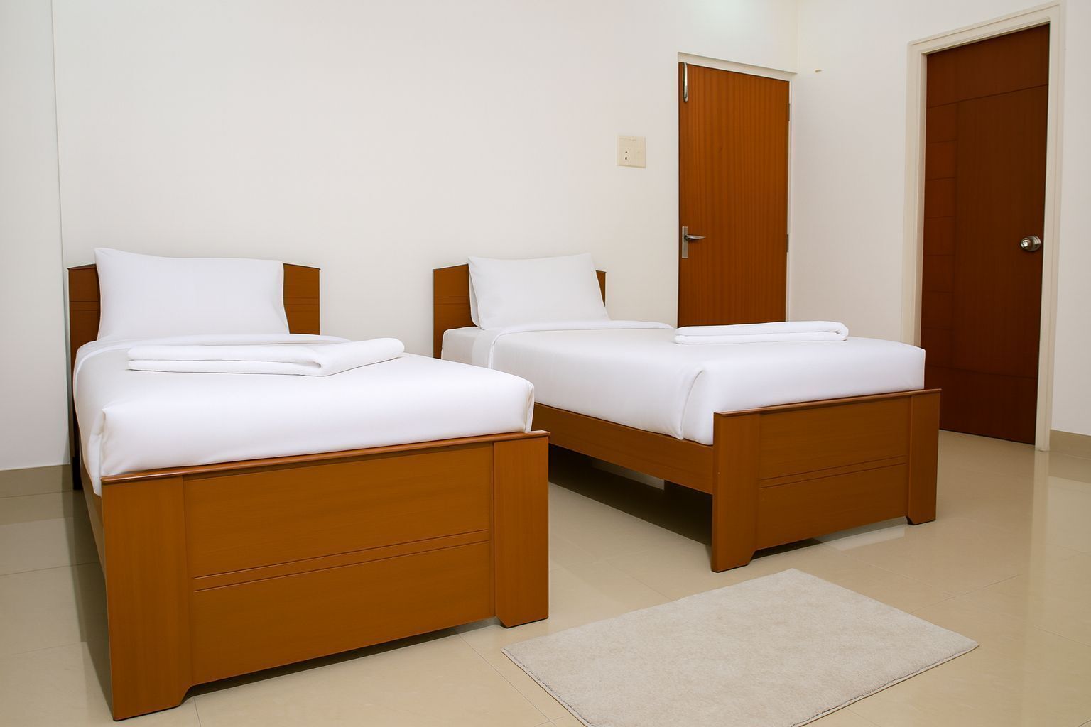 Una habitación con dos camas de madera, cada una con ropa de cama blanca y un cojín a juego. Hay una puerta de madera y un interruptor de luz en la pared.