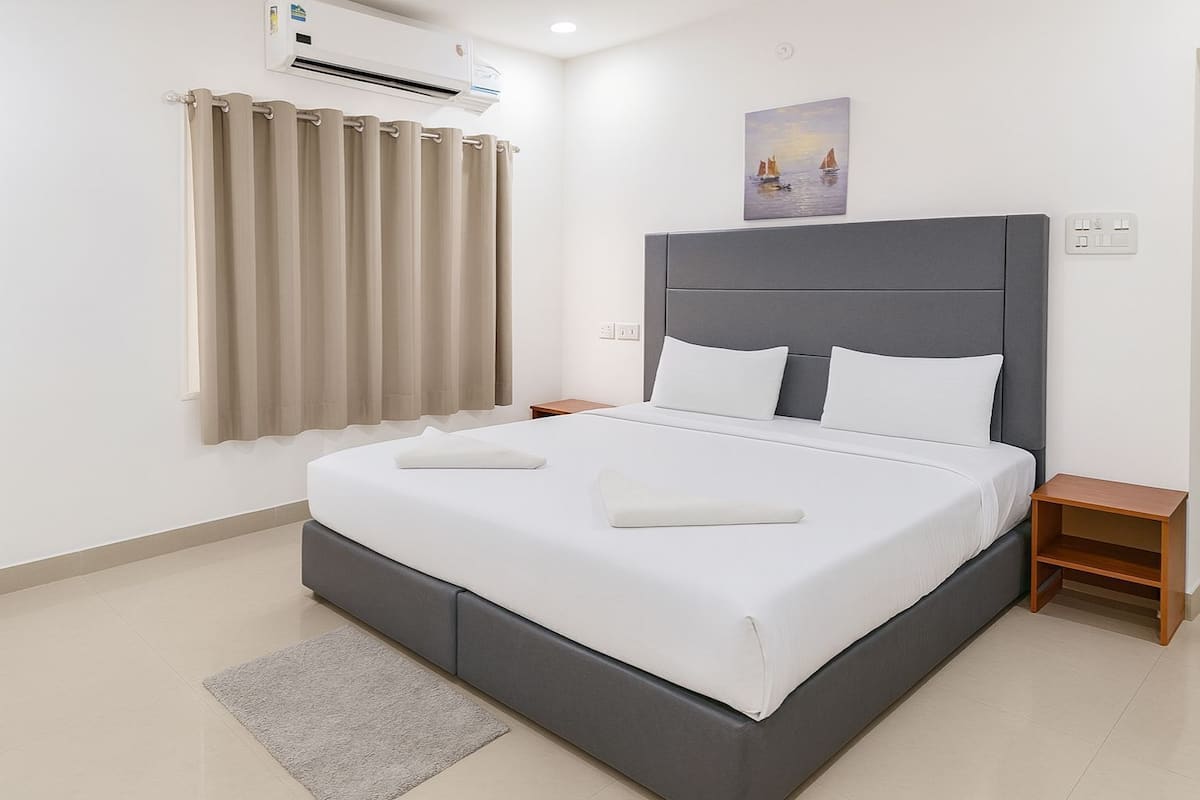 Ein modernes Schlafzimmer mit einem großen Bett, einem grauen Kopfteil, weißer Bettwäsche, einem grauen Teppich, einem kleinen Nachttisch aus Holz und einem Bild an der Wand.