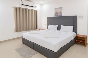 A modern bedroom with a large bed, grey headboard, white bedding, a grey rug, a small wooden nightstand, and a painting on the wall.