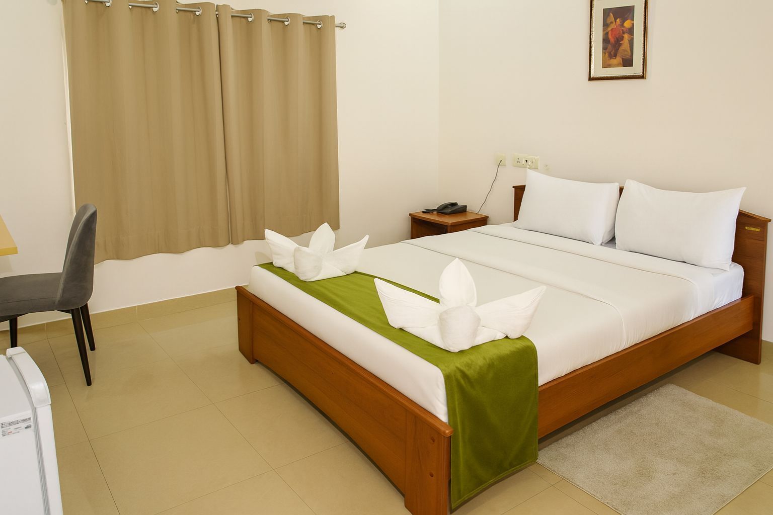 A neatly made bed with white linens and a green accent, a bedside table with a phone, a chair, and a curtain.