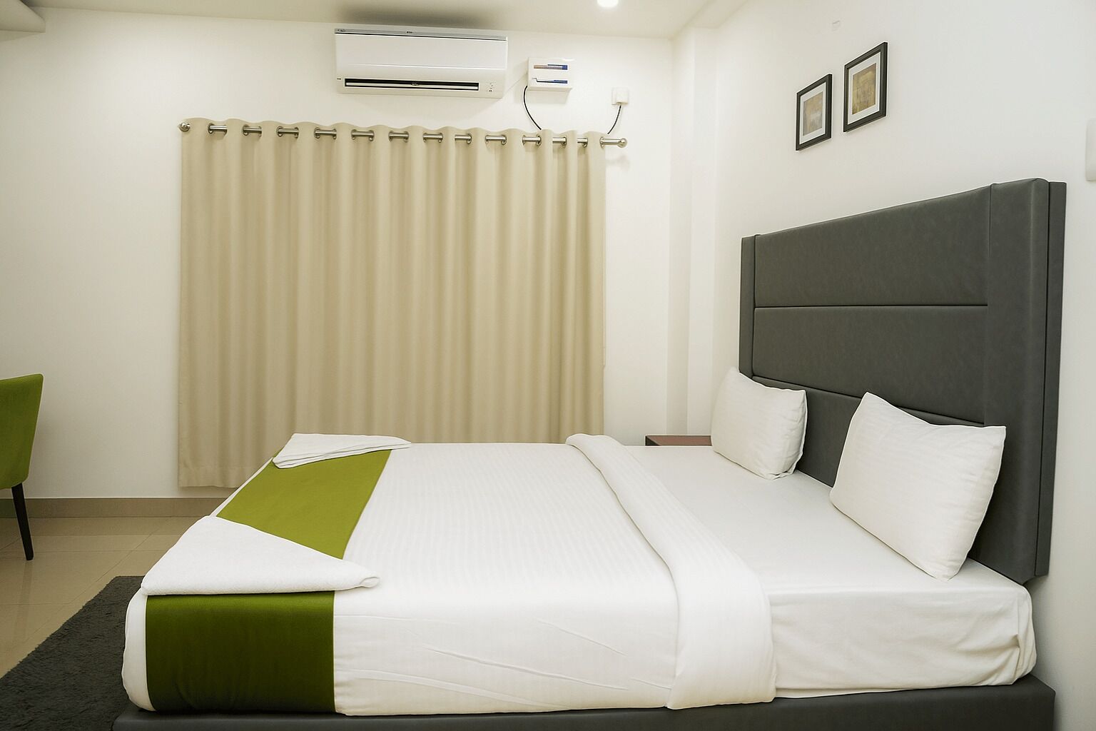 A hotel room with a bed, a green chair, a window with curtains, and an air conditioning unit.
