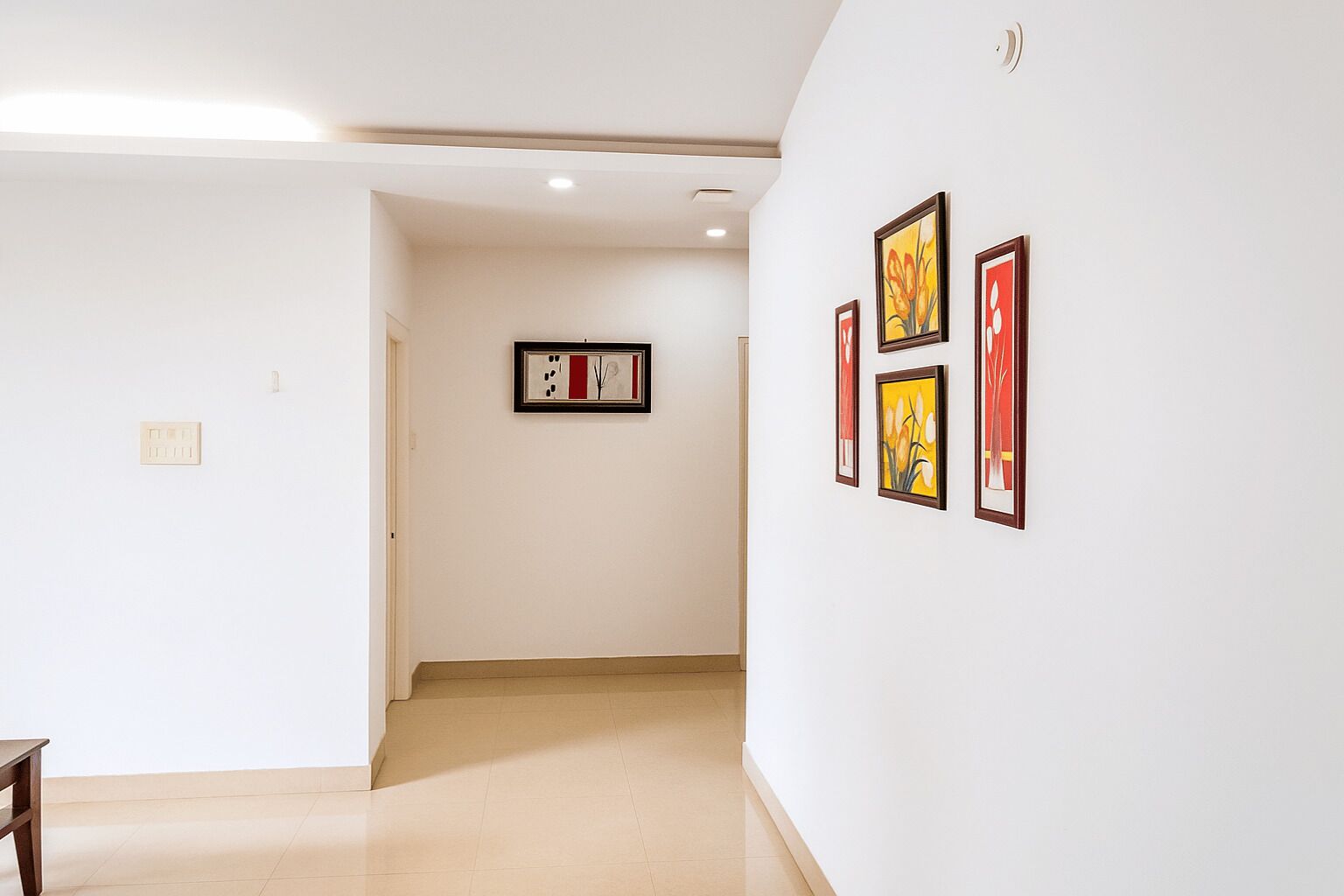 Un couloir avec un plancher en bois, des murs blancs et une porte au fond. Quatre œuvres d’art encadrées sont accrochées au mur de droite.
