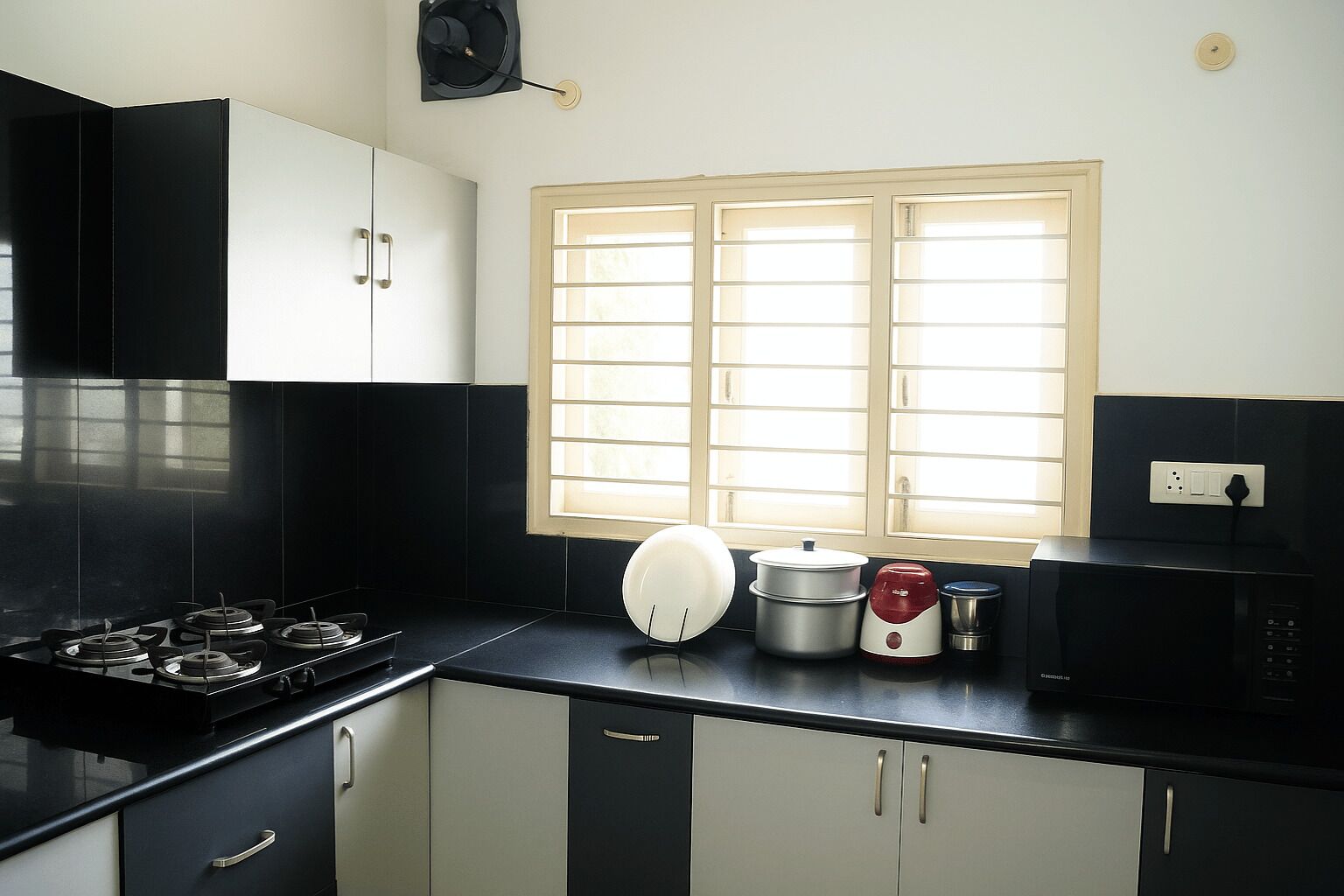 A kitchen with black cabinets, a gas stove, a microwave, a kettle, and a window with blinds.