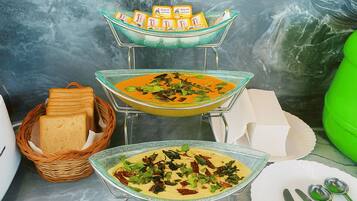 Un buffet con tre ciotole di vetro con zuppe, un cestino di pane e un contenitore verde.