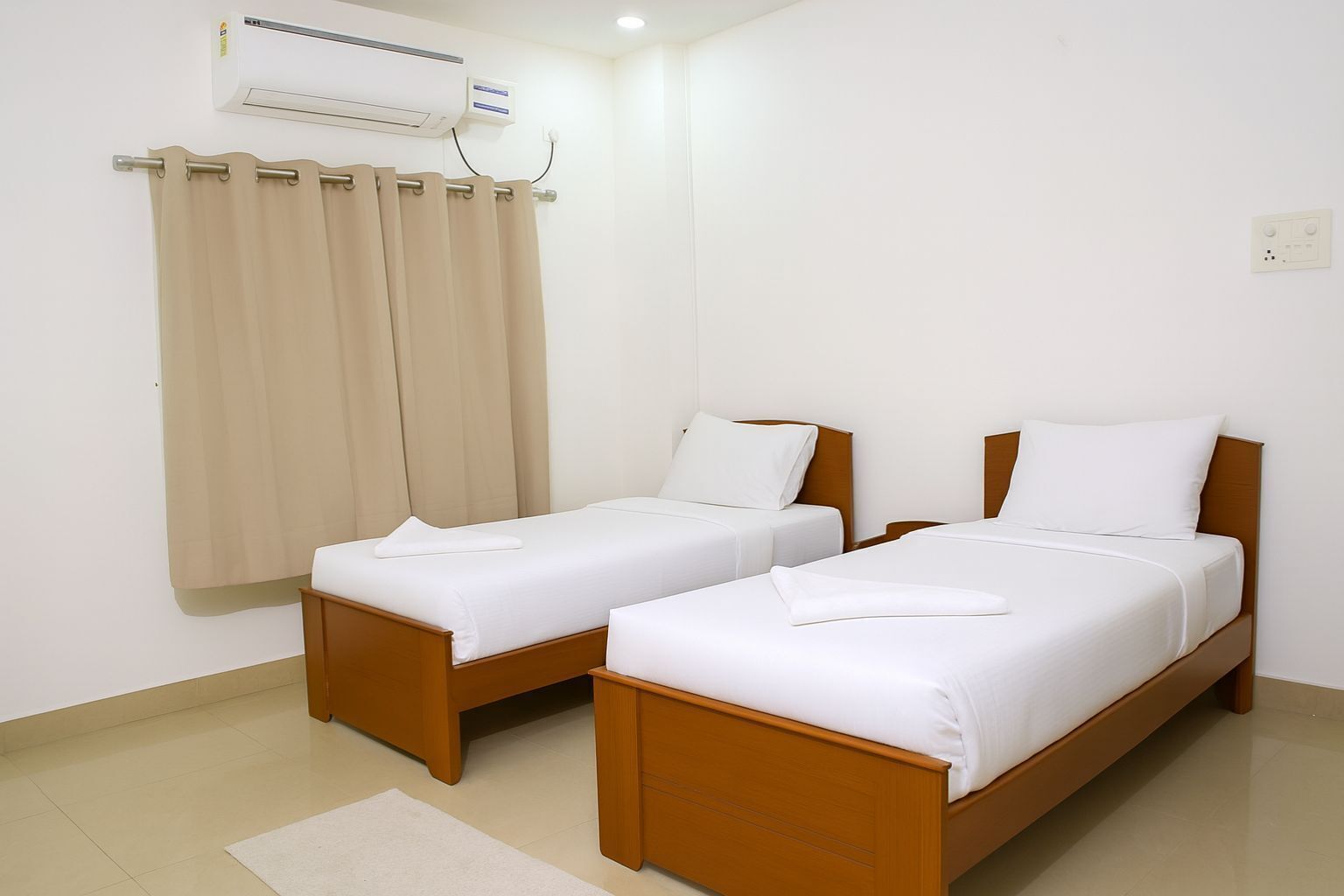 Une chambre d’hôtel avec deux lits simples, un rideau beige, un appareil de climatisation et un sol blanc.