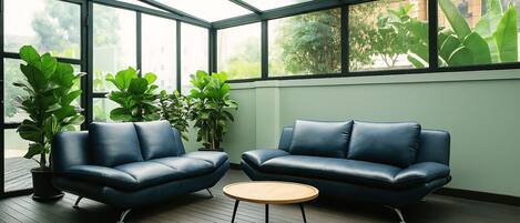 A modern room with two leather sofas, a round wooden table, and large windows with plants.