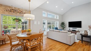 A spacious living room with a dining area, large windows, a flat-screen TV, and a stone accent wall. - NEW DECK!  Private 1 Acre Family Friendly Home - Heated Pool, Home Office & Gym (Westhampton)