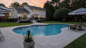 A swimming pool with a curved edge, surrounded by a paved patio. There are lounge chairs and umbrellas, and a house with a deck in the background. - NEW DECK!  Private 1 Acre Family Friendly Home - Heated Pool, Home Office & Gym (Westhampton)