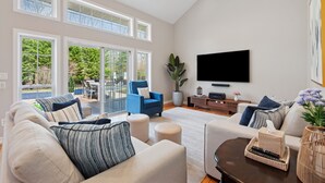 A spacious living room with a large TV, comfortable seating, and a sliding glass door leading to an outdoor area. - NEW DECK!  Private 1 Acre Family Friendly Home - Heated Pool, Home Office & Gym (Westhampton)