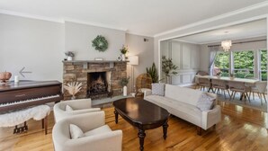 A spacious living room with a piano, a fireplace, a dining area, and large windows offering a view of greenery. - NEW DECK!  Private 1 Acre Family Friendly Home - Heated Pool, Home Office & Gym (Westhampton)