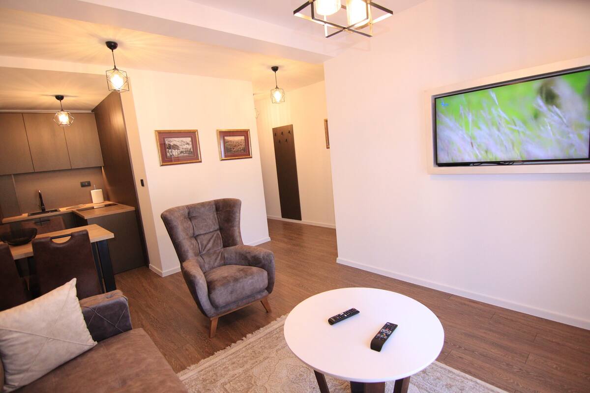 Ein modernes Wohnzimmer mit einer Couch, einem Sessel und einem runden Tisch mit zwei Fernbedienungen. Ein Flachbildfernseher ist an der Wand montiert.