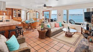 A spacious living area with a large window overlooking a body of water, a kitchen with wooden cabinets, and a dining area with wicker furniture. - Best View in Mission Bay, California (San Diego)