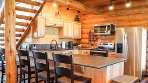 A kitchen with wooden beams, a central island with bar stools, stainless steel appliances, and light-colored cabinets. - Hill Cabin Hideaway, Romantic! Country Cabin for 2 (North Powder)