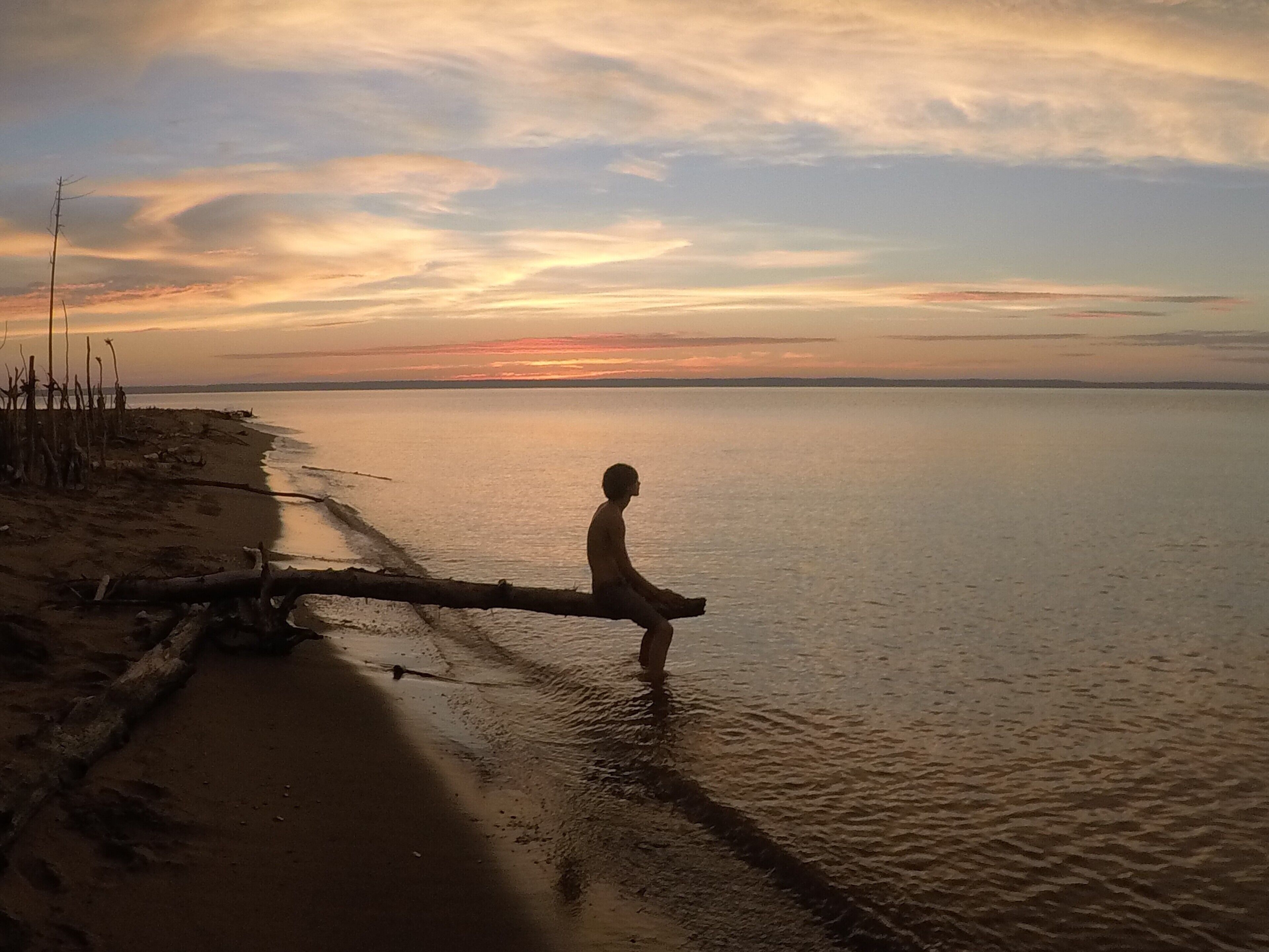 A beach scene with a person sitting on a log by the water during sunset.
