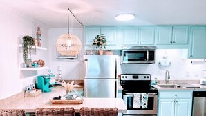 Cocina moderna con gabinetes color turquesa, un refrigerador de acero inoxidable, un microondas y una estufa. Hay sillas de mimbre y un lámpara colgante.