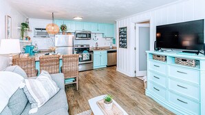Una cocina moderna con armarios color turquesa, un televisor grande, muebles de mimbre y una sala de estar acogedora con sofá y cojines.