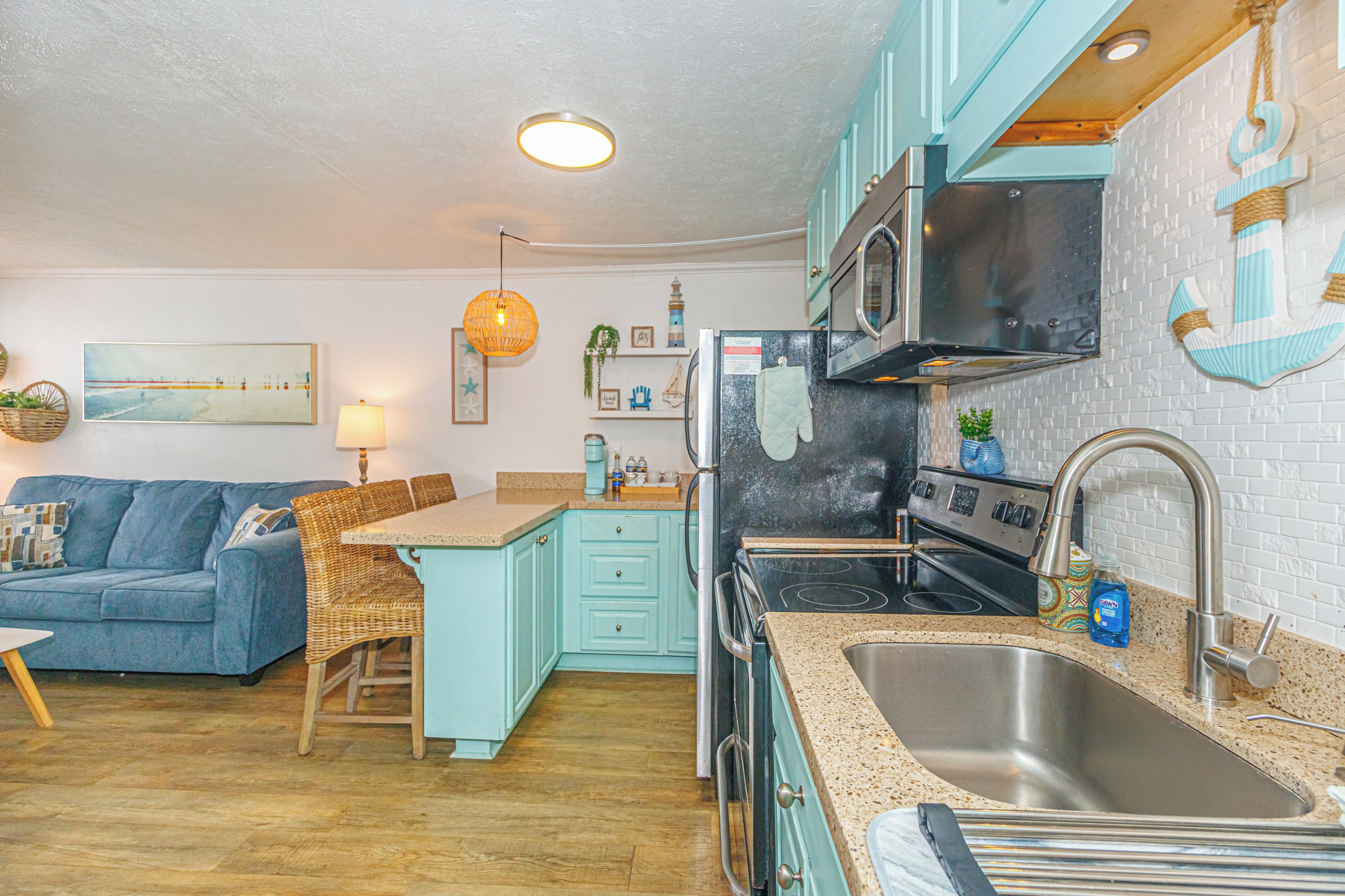 A compact kitchen with a microwave, stove, and sink. There is a blue sofa and a wicker chair in the living area.