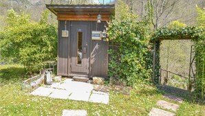Una pequeña cabaña de madera con puerta y ventanas, rodeada de follaje y un sendero de piedra.