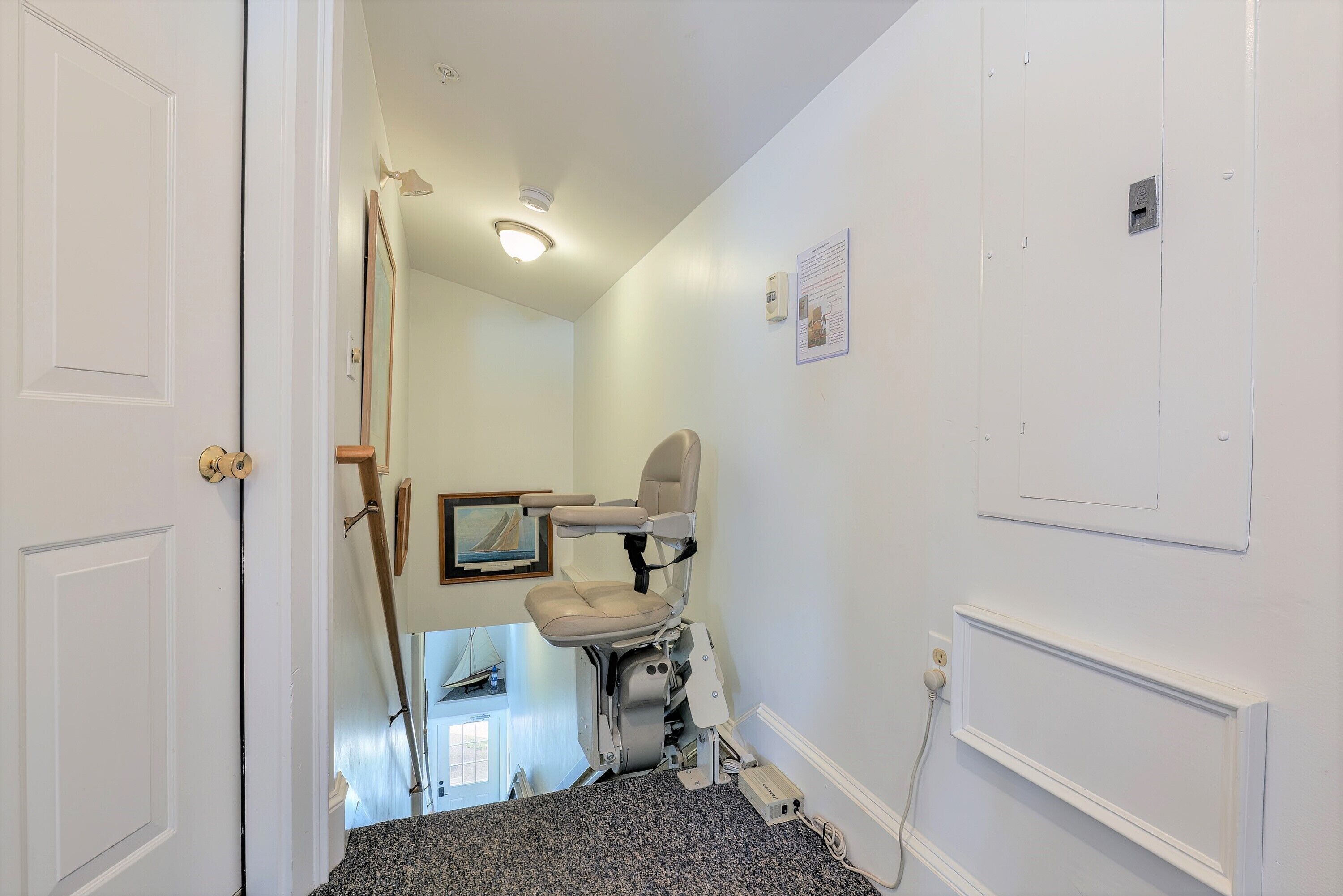 Un couloir étroit avec une porte blanche sur la gauche, un fauteuil roulant au centre et un mur blanc orné d’une petite boîte électrique et d’un tableau encadré.
