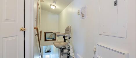 Un couloir étroit avec une porte blanche sur la gauche, un fauteuil roulant au centre et un mur blanc orné d’une petite boîte électrique et d’un tableau encadré.