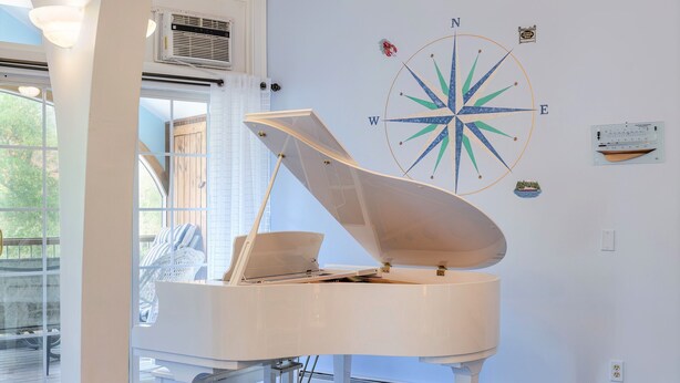 Un piano à queue dans une pièce décorée d’un motif de boussole sur un mur, avec une fenêtre munie de stores et un ventilateur de plafond.