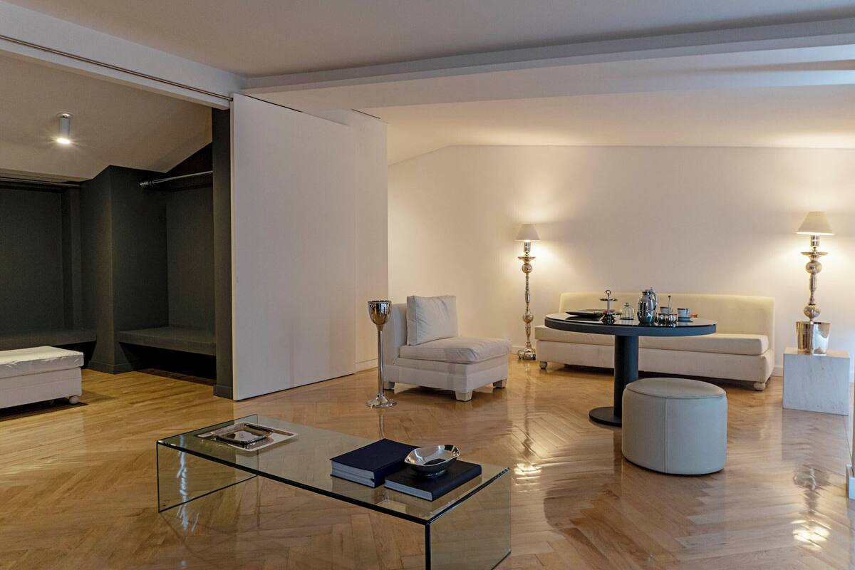 A modern living room with a glass coffee table, white sofas, a round table with decorative items, and a staircase leading to a dark room.
