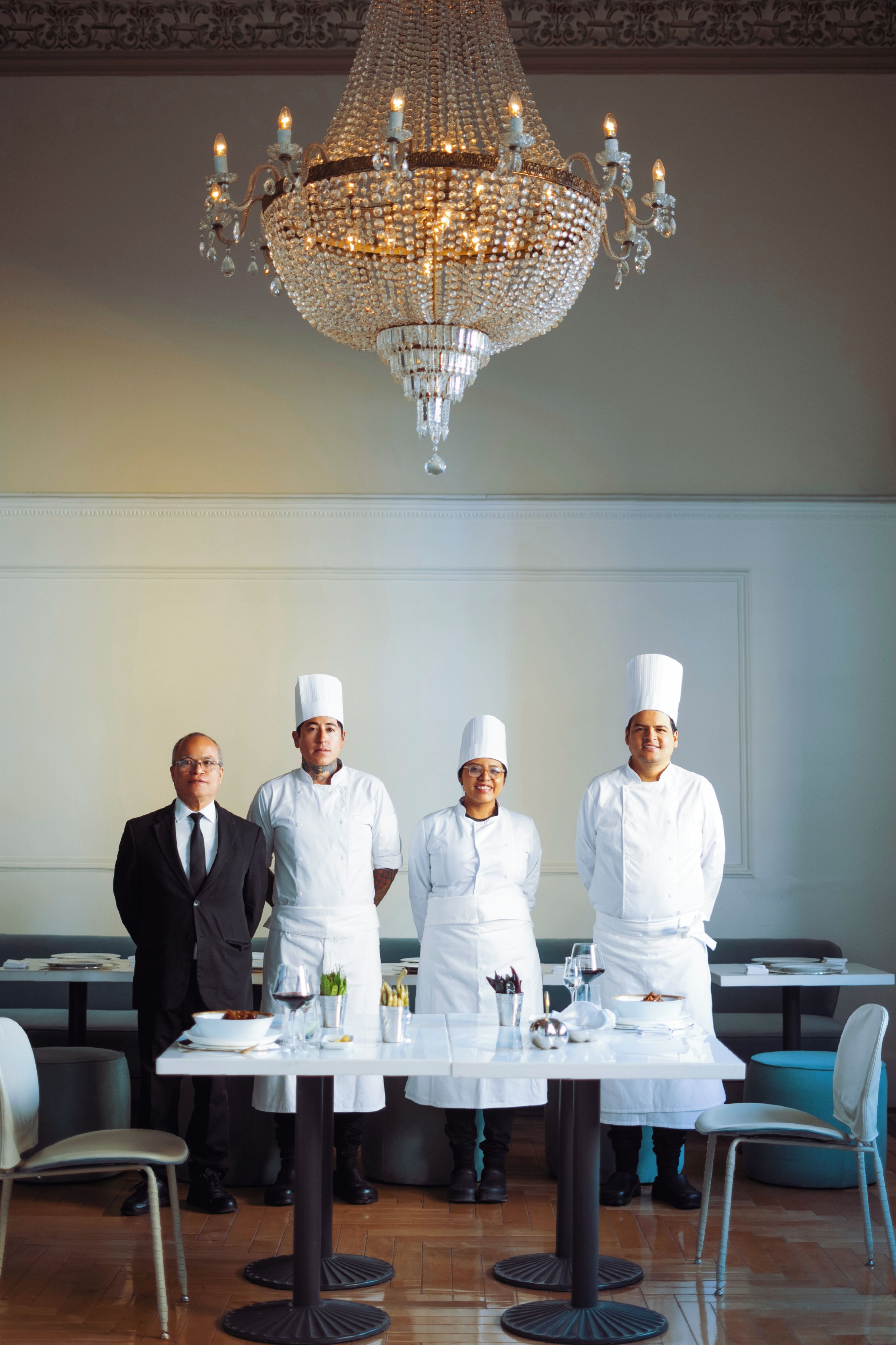 Un elegante comedor con candelabro, mesa dispuesta para cuatro personas y un camarero con uniforme.