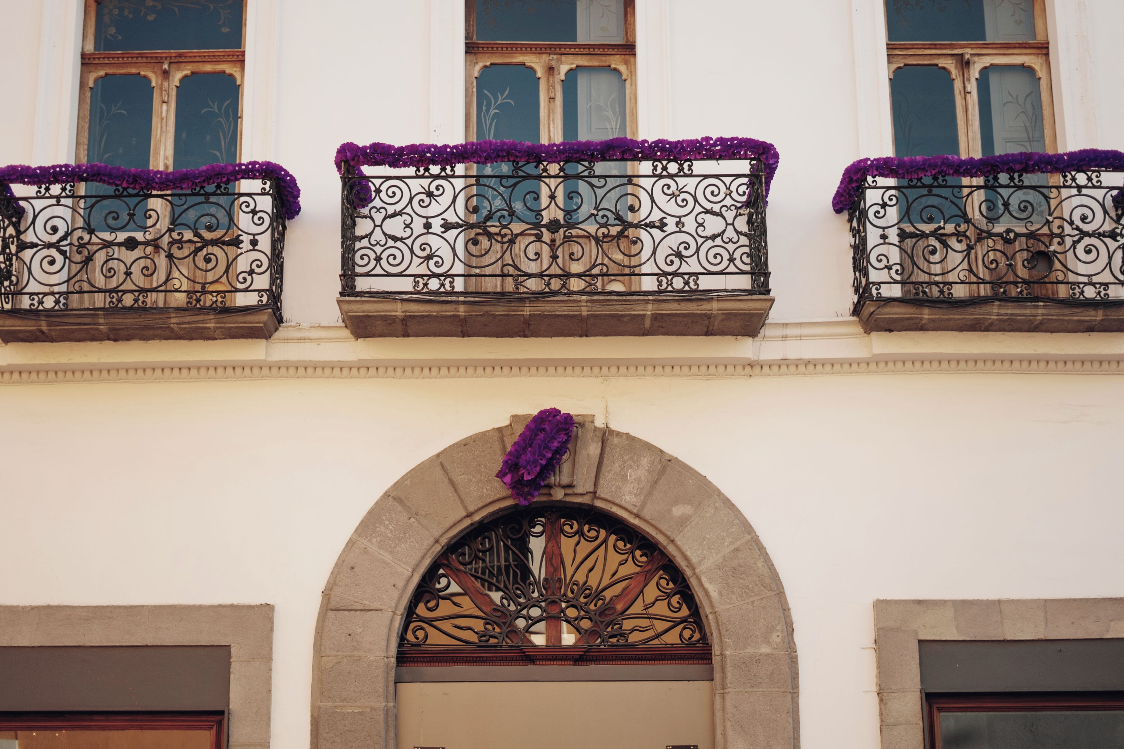 Une façade d’immeuble ornée de balcons en fer forgé ouvragé, décorés de fleurs violettes, avec une entrée centrale en arc de cercle et un portail en métal décoratif.