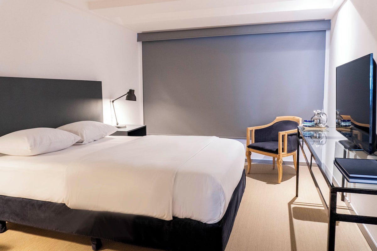 A hotel room with a bed, a desk, a chair, a lamp, and a television.